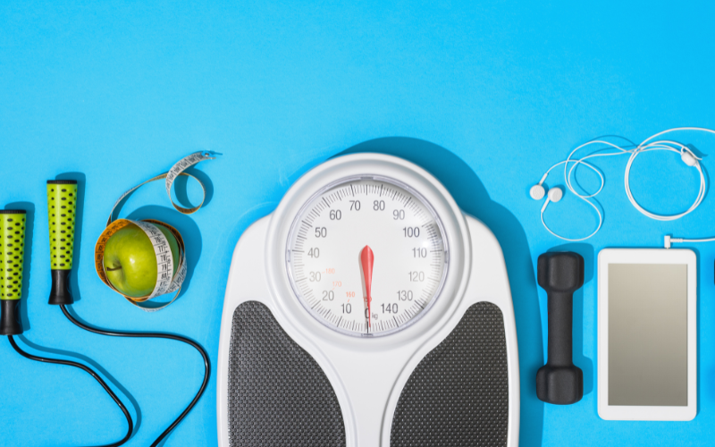 Weight scale next to a jump rope, apple, tape measure, dumbbell, and mobile phone