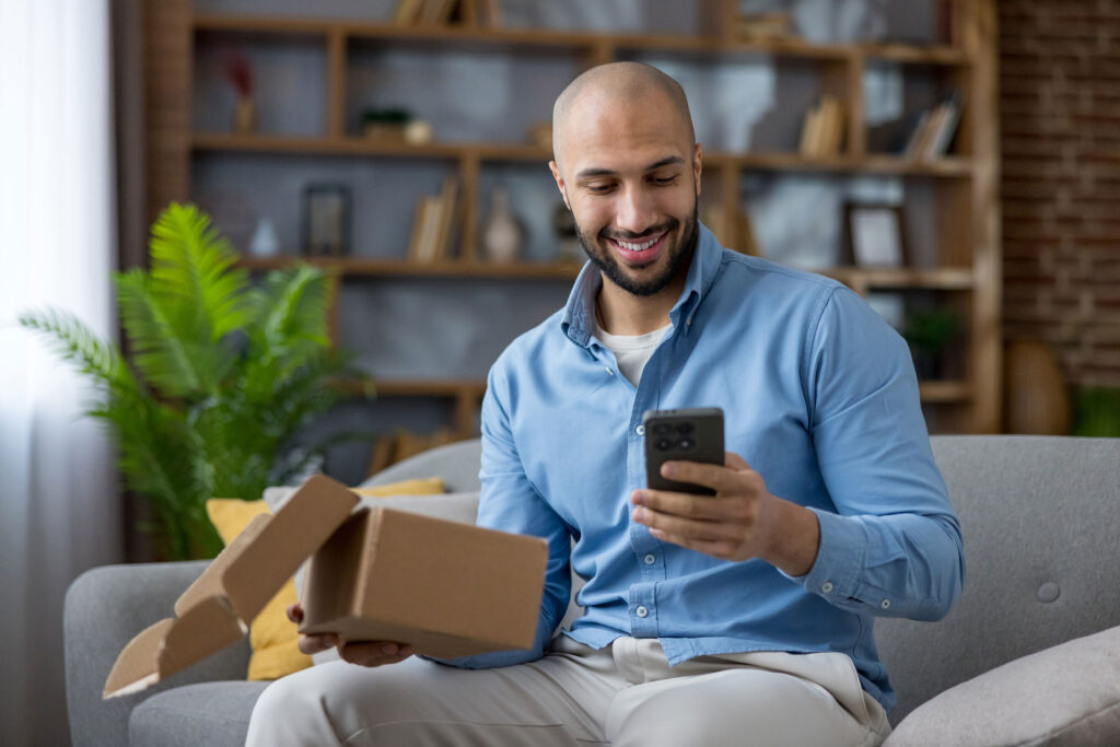 Smiling man unboxing a cardboard package on his couch at home while checking his smartphone after online shopping, relaxed and satisfied with his new purchase and delivery
